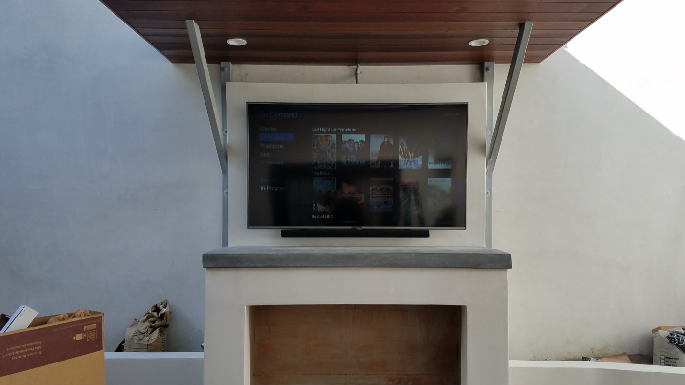 TV mounted outdoors on a stucco fireplace under a wood pergola