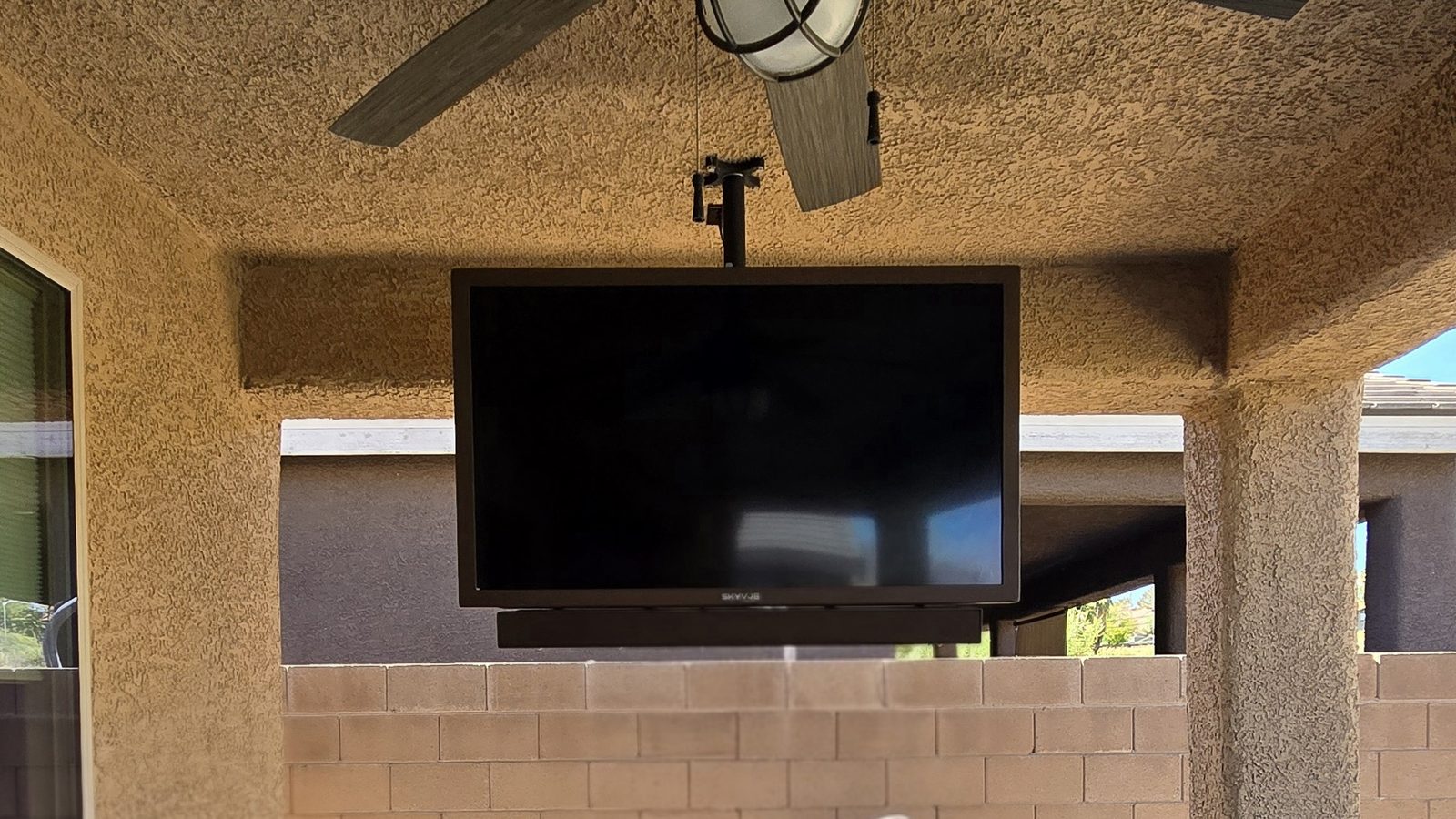 Outdoor TV mounted on a stucco covered patio wall directly beneath an outdoor ceiling fan, with a sound bar below and a block wall behind